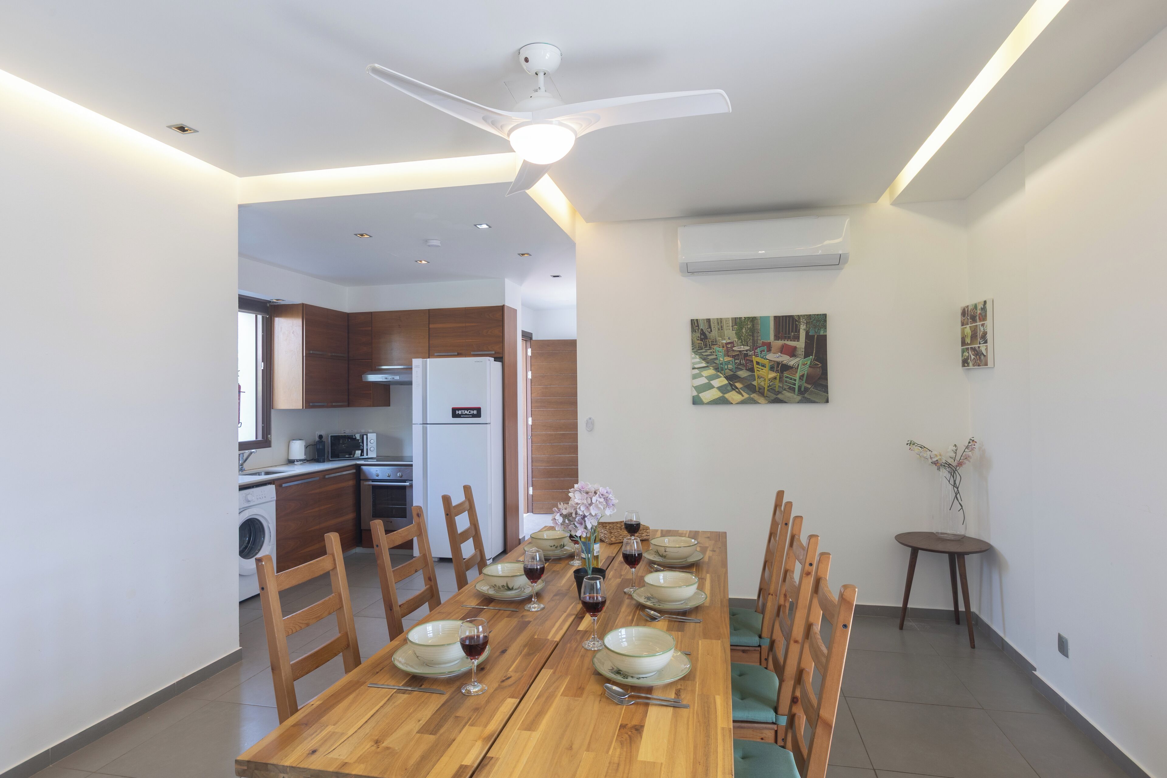 Uma sala de jantar com mesa de madeira posta para seis pessoas, rodeada de cadeiras, e um ventilador de teto acima.