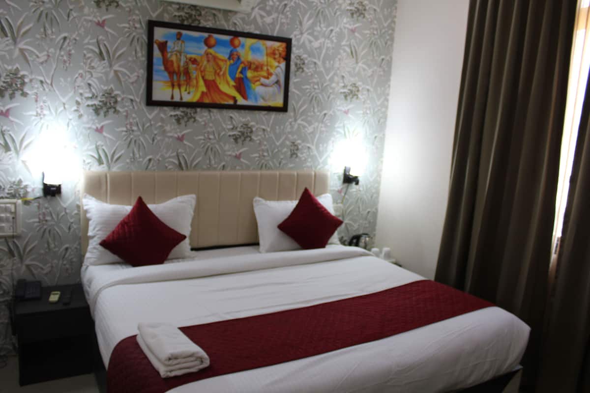 Quarto de hotel com uma cama grande, duas almofadas vermelhas, um edredom branco e um cobertor vermelho. Há uma pintura na parede e uma mesa de cabeceira com um telefone e um abajur.