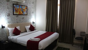 A hotel room with a large bed, a nightstand, a desk, and a chair. - Avexia Premiere (Chittorgarh)
