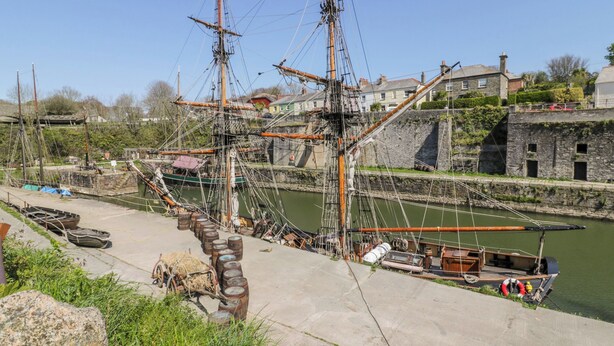 A historic ship docked at a harbor with a stone wall and buildings in the background. - Bumblebee Cottage (Bodmin)