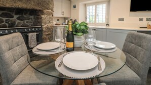 Zona pranzo con tavolo in vetro, sedie rivestite in grigio, parete in pietra, cucina sullo sfondo e televisore a schermo piatto montato a parete.