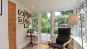 A room with a wooden floor, a black cushioned chair, a small table with a plant, and a large window with a view of greenery. - Woodman Cottage (Morecambe)