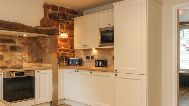 A kitchen with white cabinets, a stone wall, a wooden floor, a built-in oven, a microwave, and a toaster. - Woodman Cottage (Morecambe)
