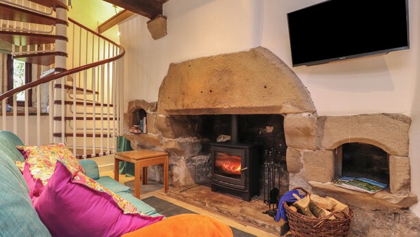A cozy living room with a stone fireplace, a flat-screen TV mounted on the wall, a wooden staircase, and a comfortable sofa with colorful cushions. - Pear Tree Cottage at Hey Farm (Barnoldswick)