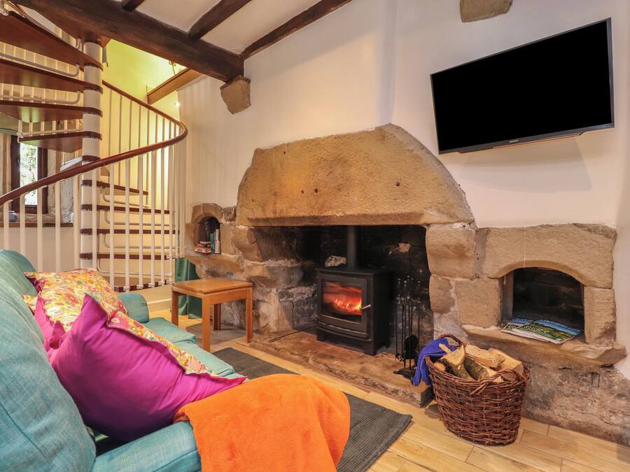 A cozy living room with a stone fireplace, a flat-screen TV mounted on the wall, a wooden staircase, and a comfortable sofa with colorful cushions.