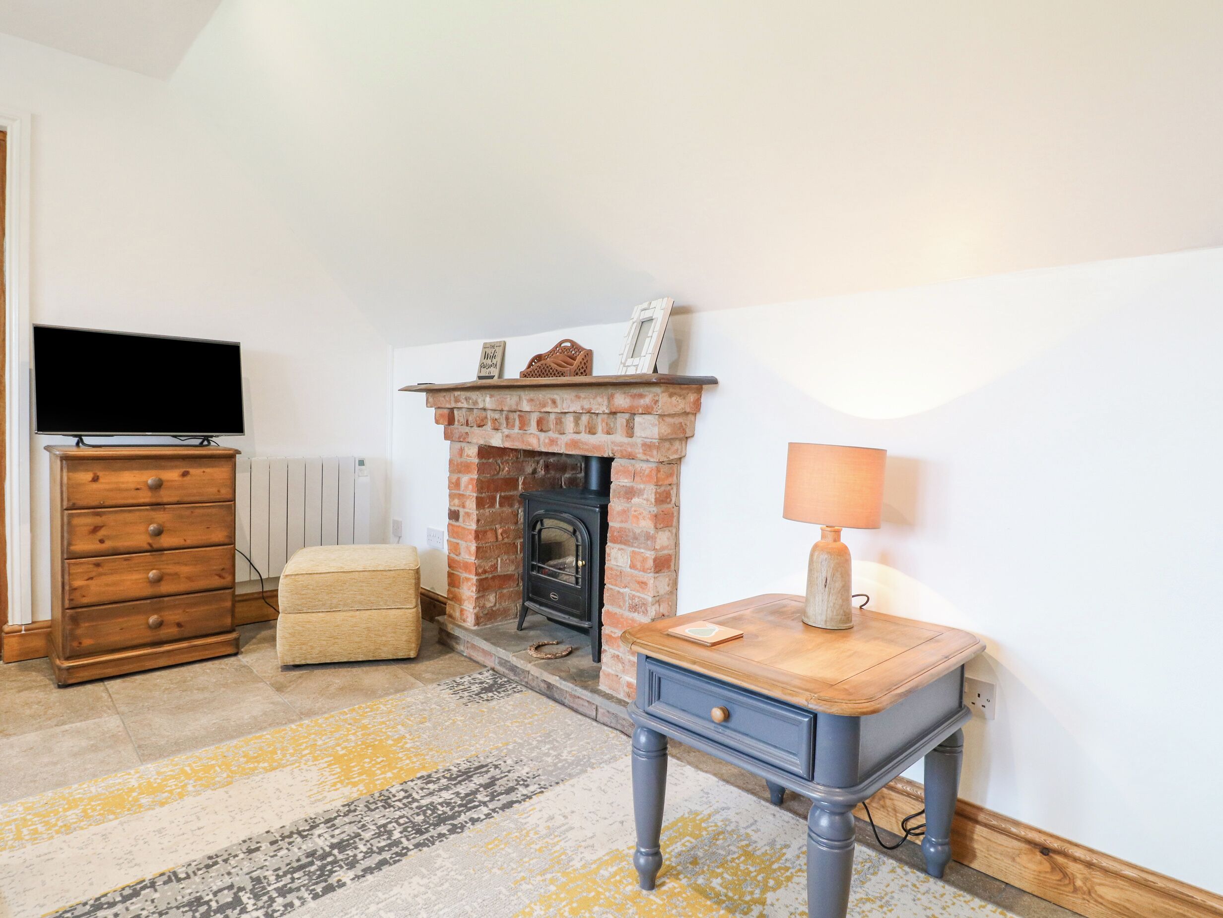 A cozy living room with a brick fireplace, a television on a wooden dresser, a small table with a lamp, and a patterned rug.