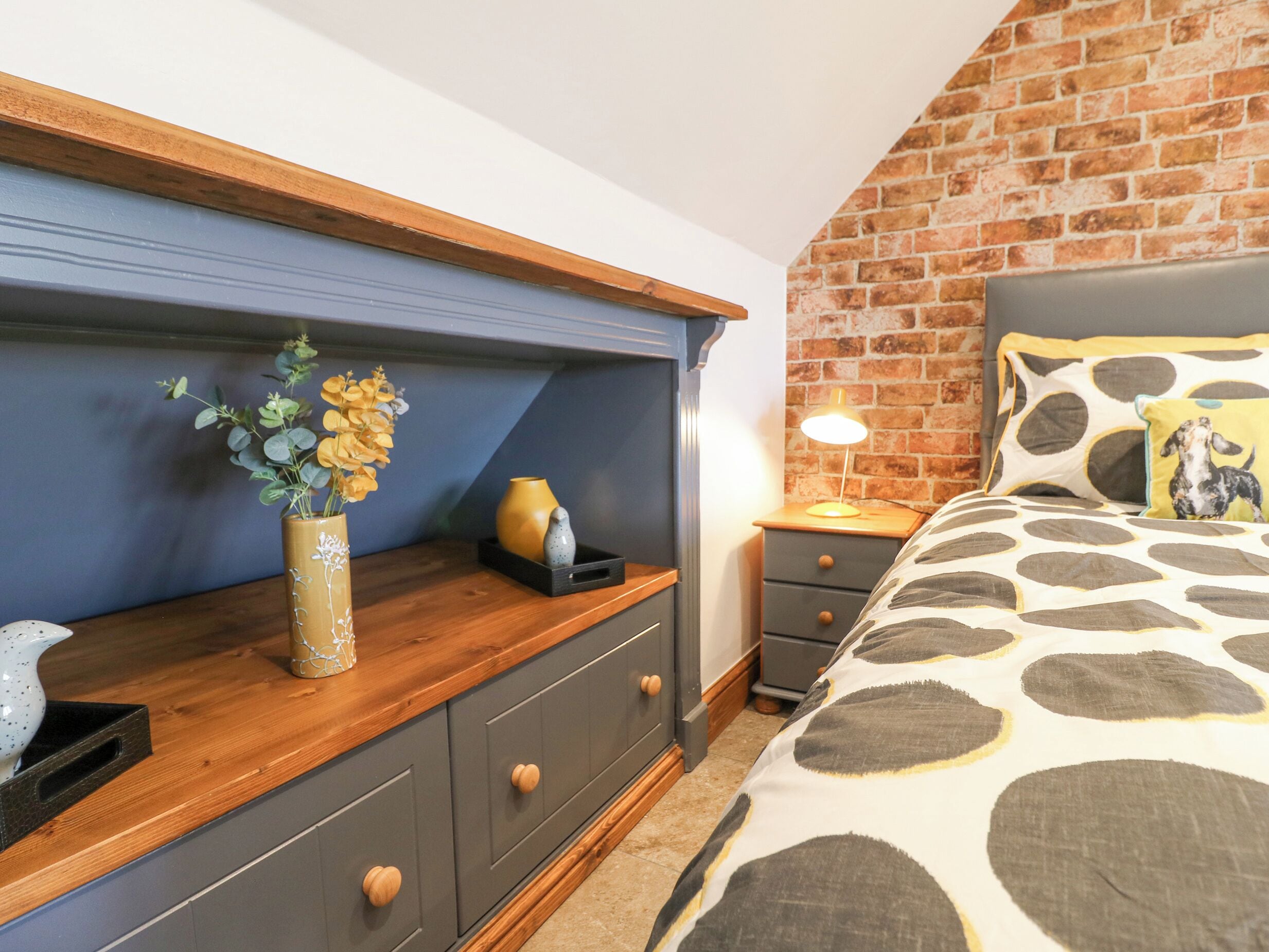 A bedroom with a brick wall, a wooden dresser, a bed with a patterned bedspread, and a nightstand with a lamp.