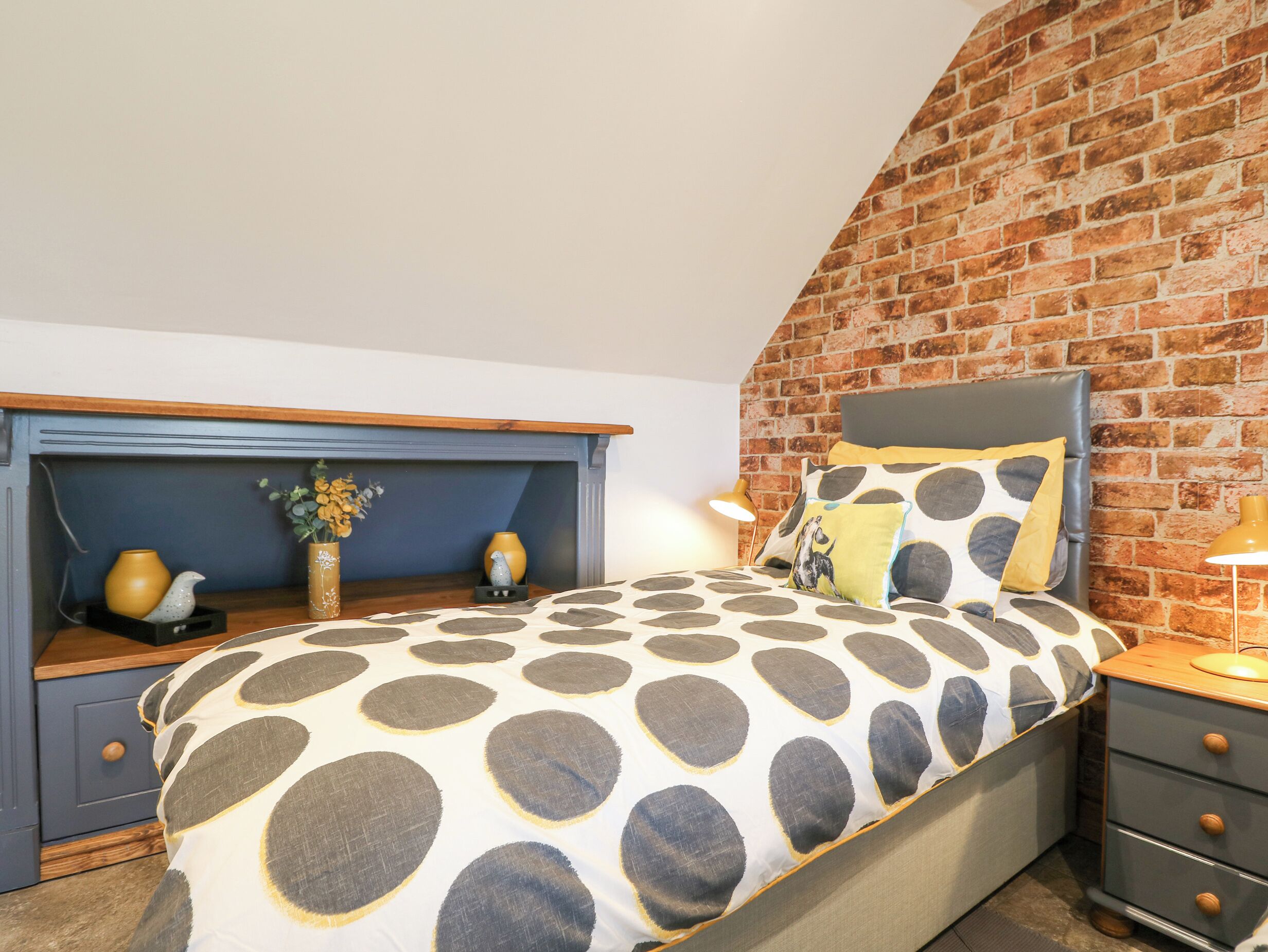 A bedroom with a brick wall, a bed with a polka dot bedspread, a nightstand, a dresser, and a small shelf with decorative items.