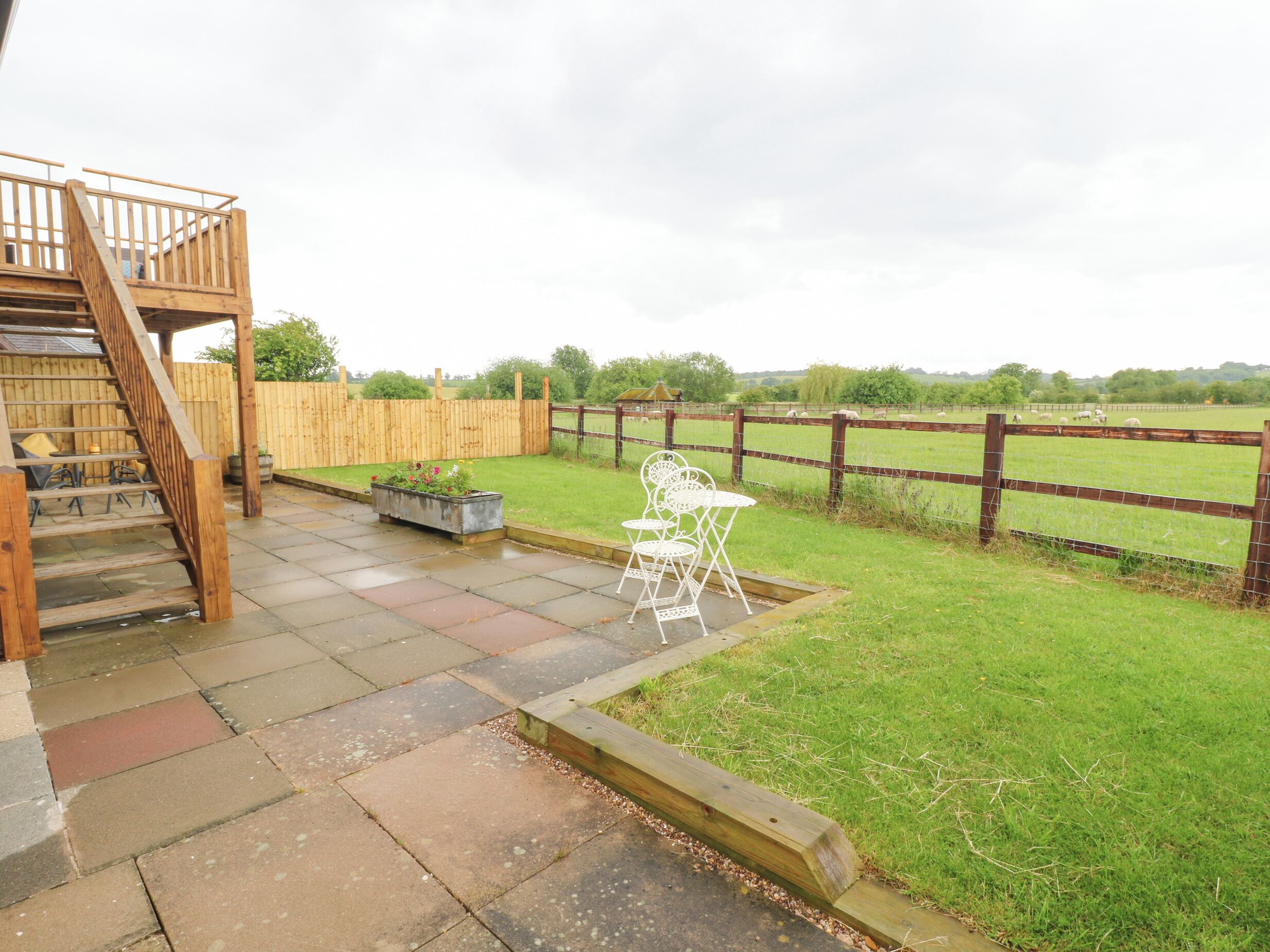 Un patio con escaleras de madera, mobiliario exterior y vista a un campo cercado.