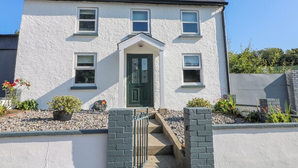 A two-story house with a green door, multiple windows, and a small front garden with plants and a gravel pathway. - An Teach Ban (Bunclody)