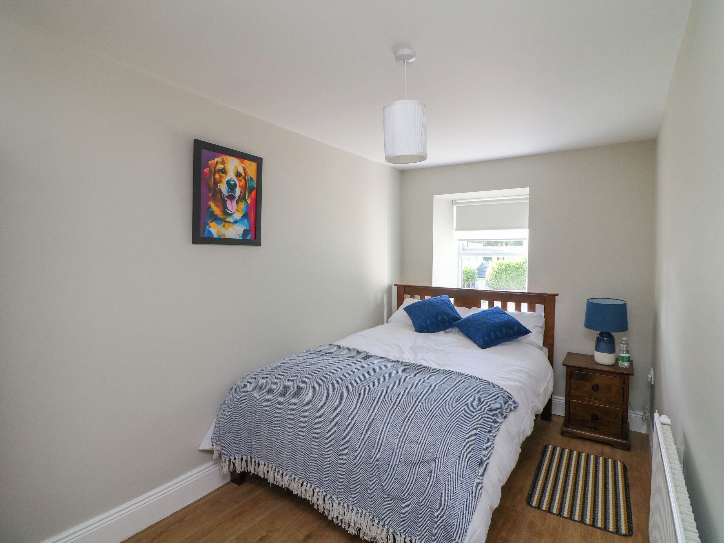A bedroom with a bed, a nightstand, a lamp, a window, and a painting on the wall.