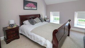 A bedroom with a wooden bed, a nightstand with a lamp, a framed picture on the wall, and two windows. - An Teach Ban (Bunclody)