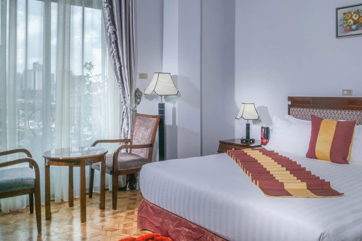 Une chambre d’hôtel comprenant un lit, deux chaises, une table, une table de chevet avec des lampes et une fenêtre avec des rideaux.