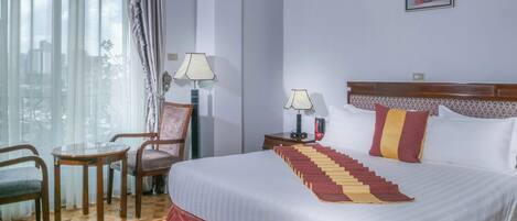Habitación de hotel con cama, dos sillones, una mesa, mesita de noche con lámparas y ventana con cortinas.