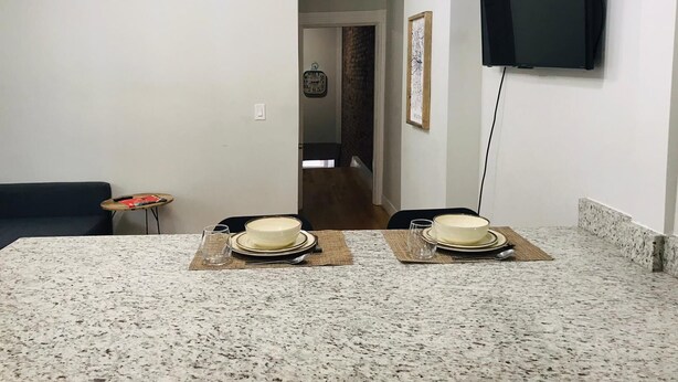 Une cuisine dotée de plans de travail en granit, une table à manger mise avec des assiettes et des bols, une petite table d’appoint et un téléviseur fixé au mur.