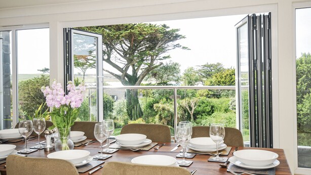 A dining area with a table set for six, large windows, and a view of greenery. - Bodare 8 (Wadebridge)