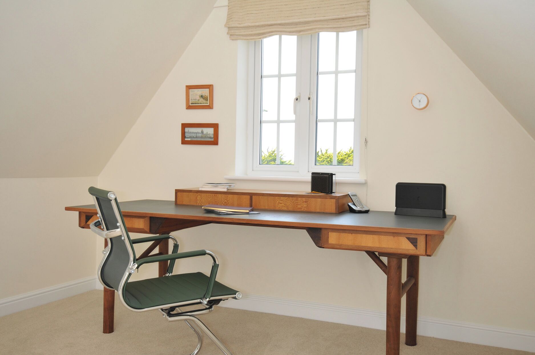 A room with a sloped ceiling, a desk with a chair, a window with blinds, and two framed pictures on the wall.