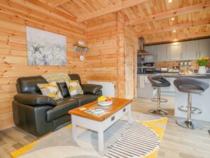 A cozy cabin interior with a wooden interior, a black leather sofa, a coffee table, and a kitchen area with modern appliances. - Little Orchard Lodge (Truro)