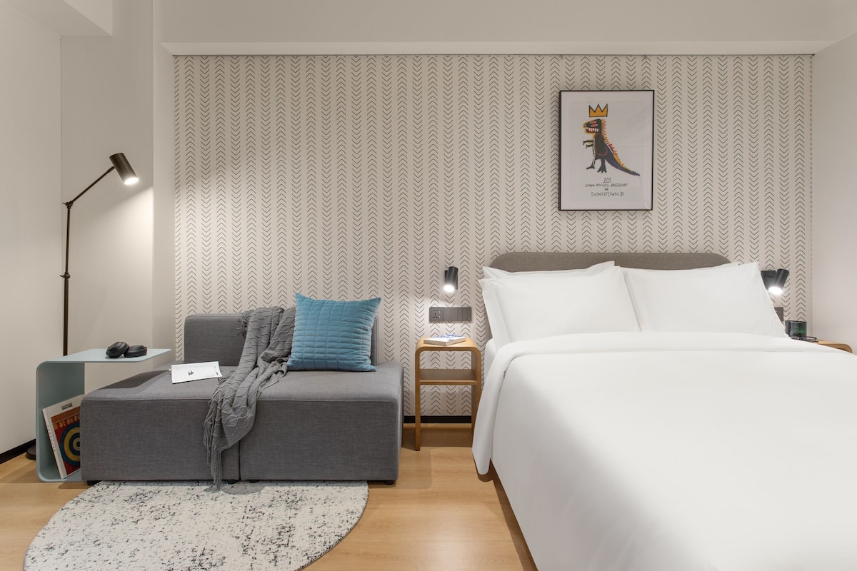 Ein modernes Hotelzimmer mit grauem Sofa, einem weißen Bett, einem kleinen Holfnachtkästchen und einem Wandbild eines Cartoon-Dinosauriers.