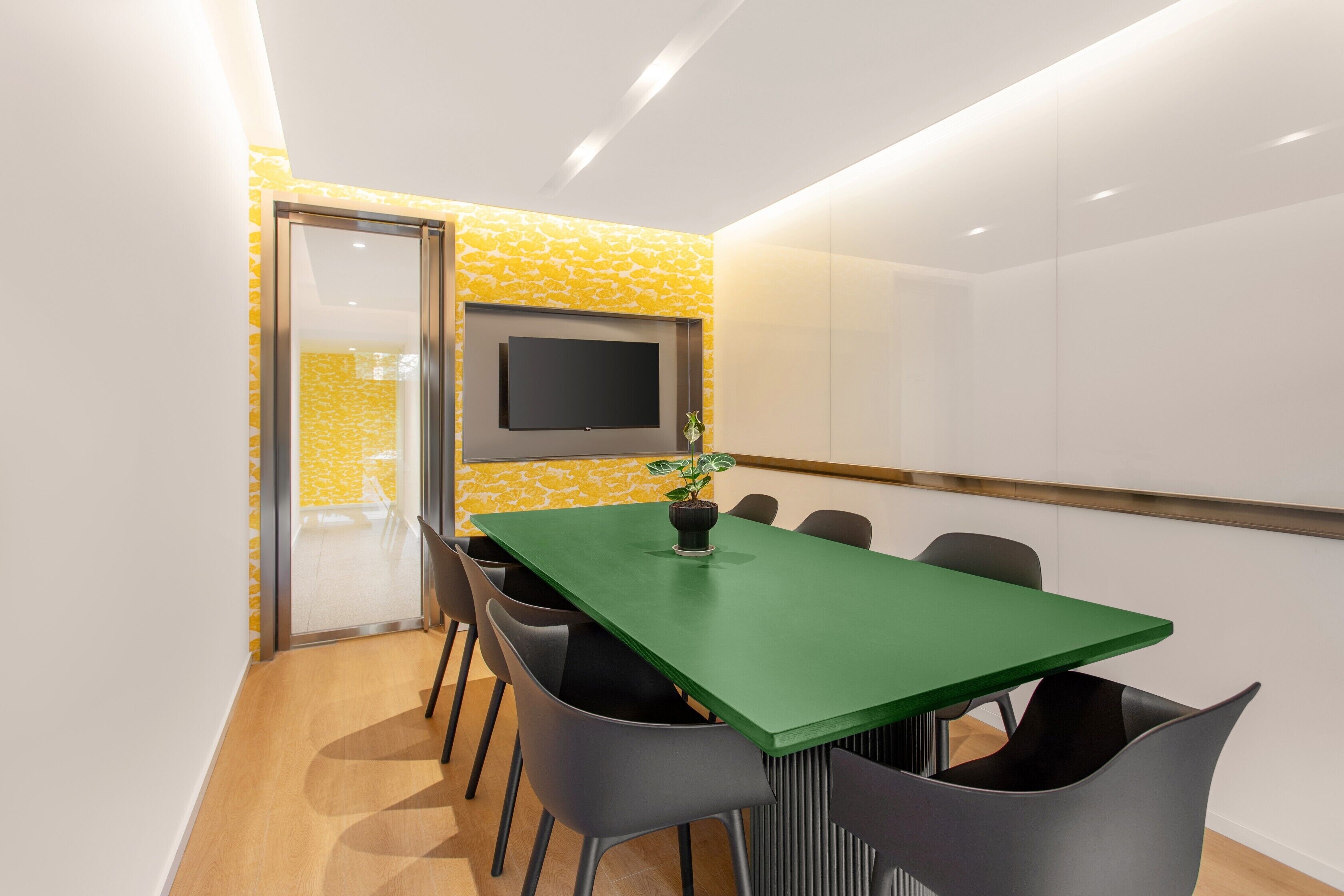 A conference room with a green table, black chairs, a potted plant, and a flat-screen TV on the wall.
