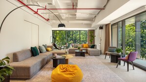 A modern living room with a sectional sofa, a yellow bean bag, and a coffee table. There are multiple chairs and a small table with a plant. The room has large windows with a view of trees outside. - Stey-Shenzhen Qianhai Hotel & Apartment (Shenzhen)