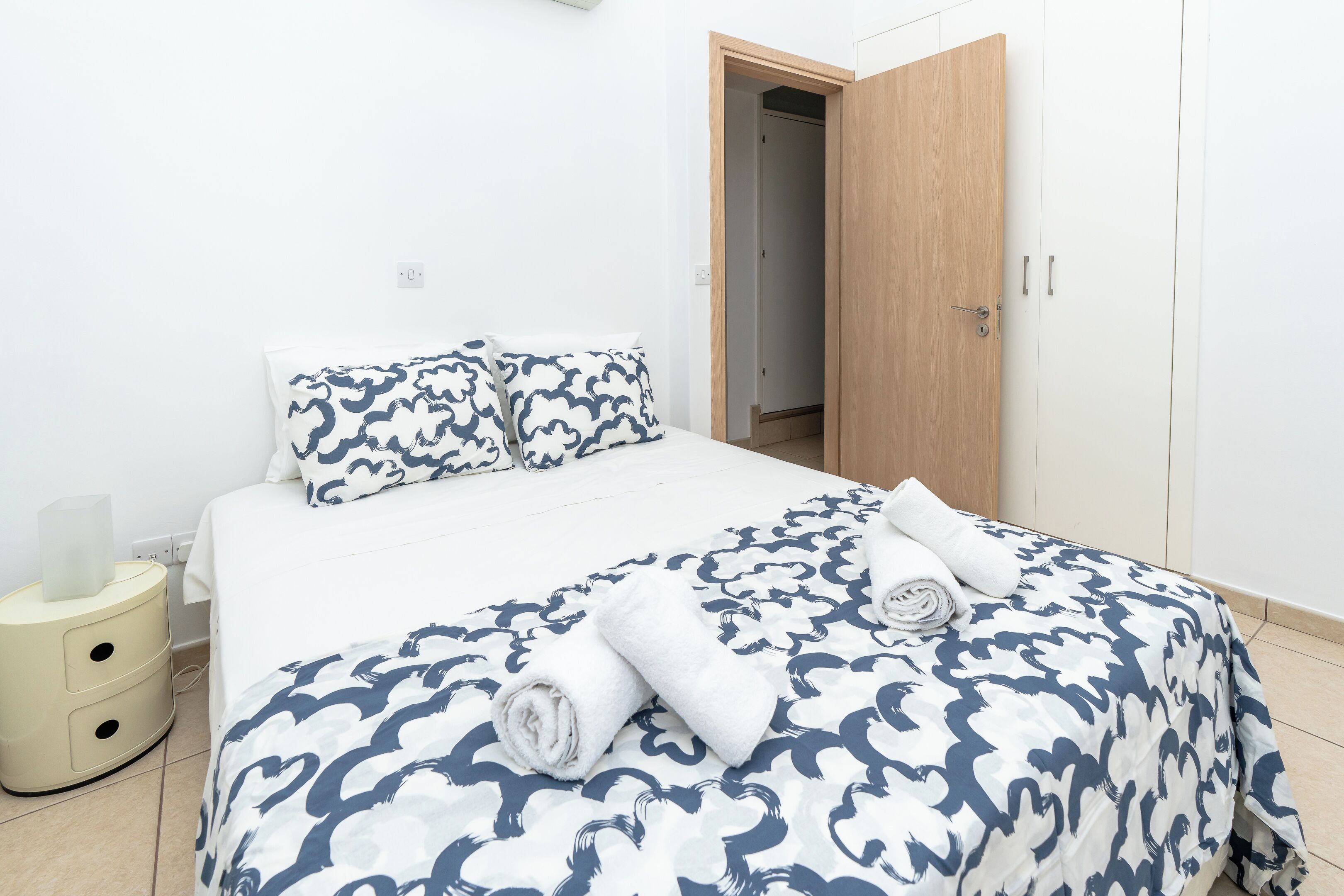 A neatly made bed with a patterned bedspread, a bedside table with a lamp, and a wooden wardrobe.