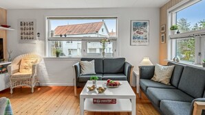 Una sala de estar moderna con un sofá, sillones y una mesa de centro. Hay ventanas con vistas a una casa y un cartel en la pared.
