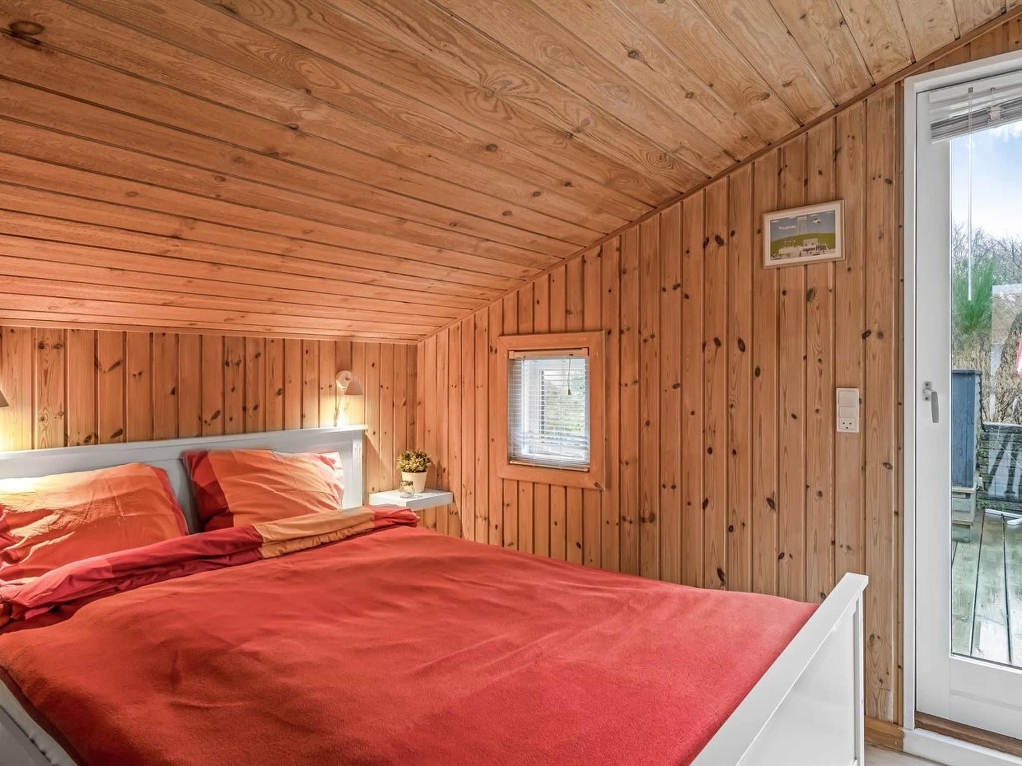 A wooden-paneled room with a bed, a window, and a small table with a lamp.