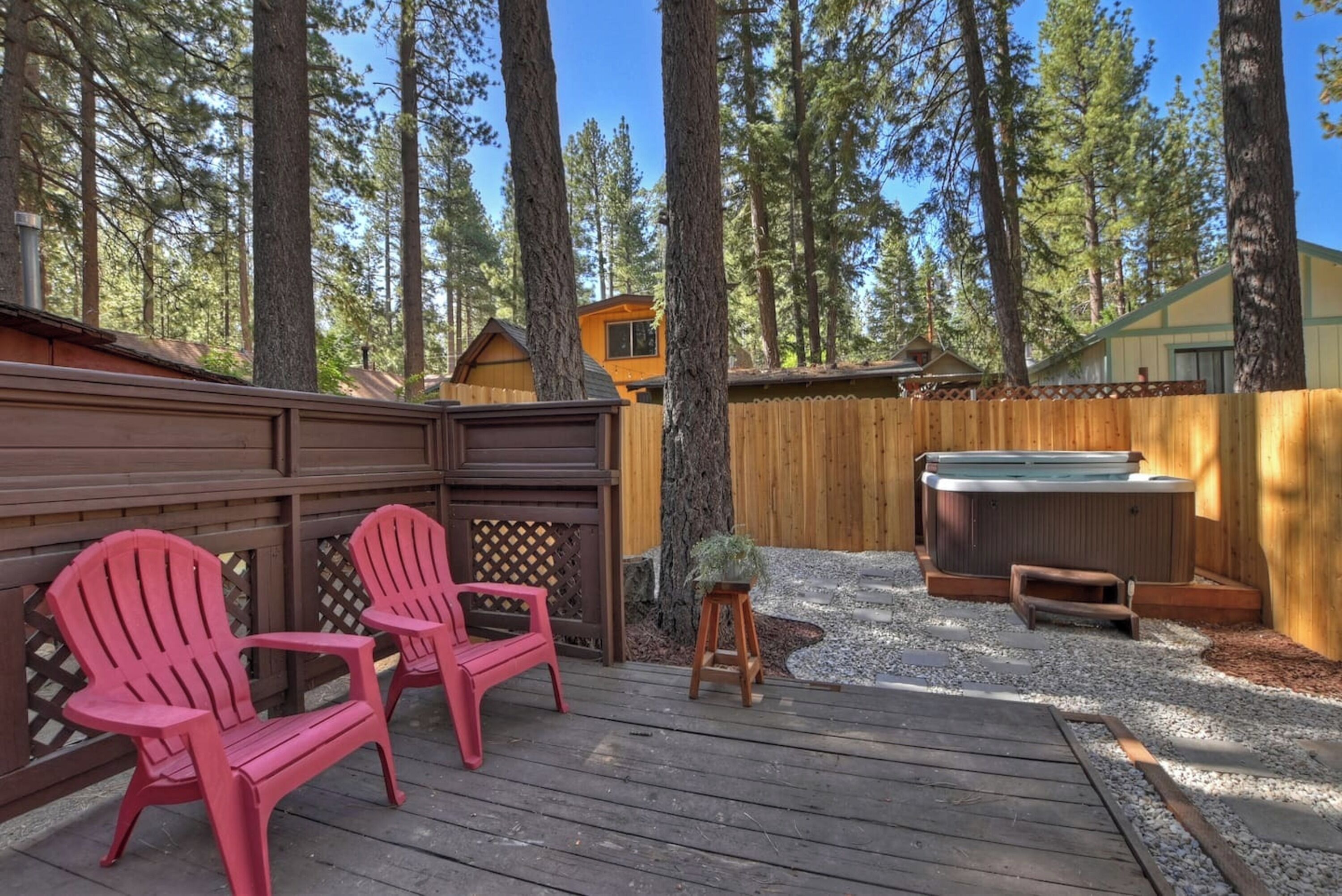 Un patio in legno con poltrone Adirondack, una vasca idromassaggio e un recinto in legno in una zona boschiva.