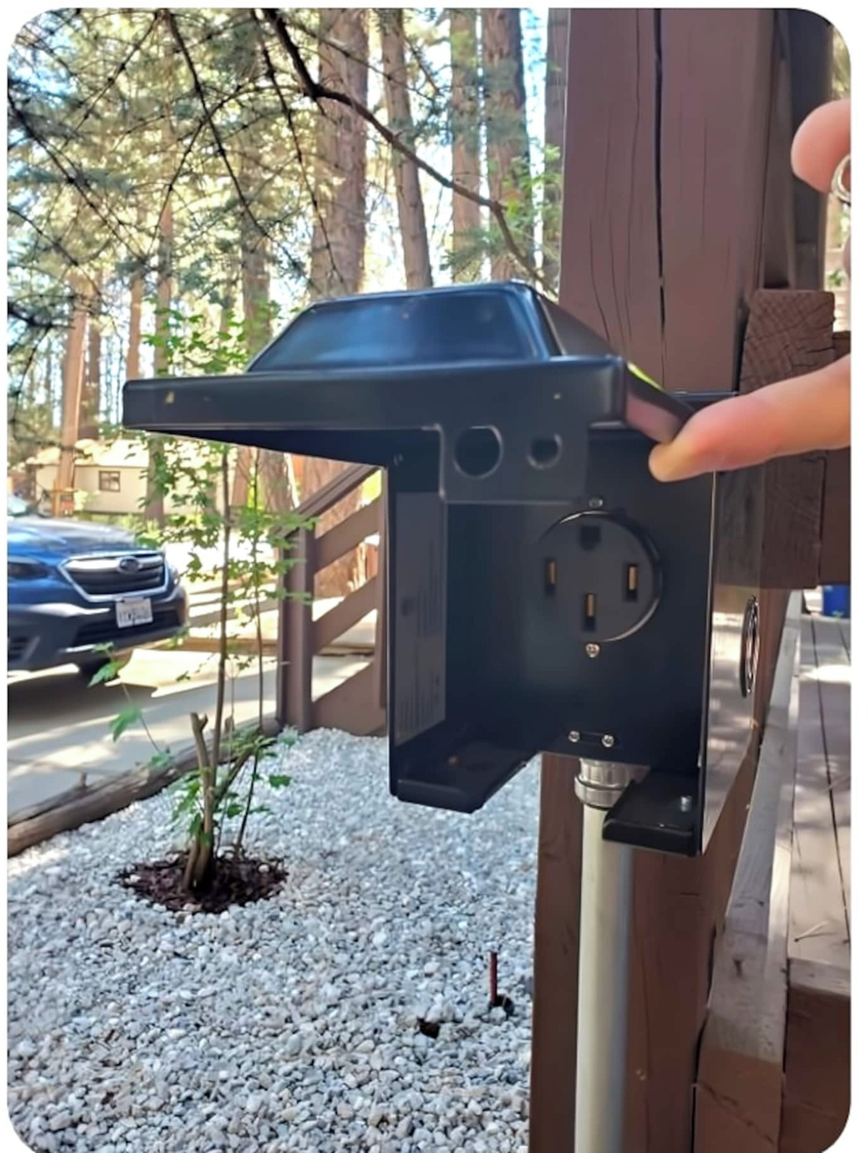 Caixa elétrica preta fixada em um poste de madeira com um mastro de metal, cercada por pedrisco e pequenas plantas.