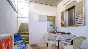 Un pequeño comedor con una mesa dispuesta para dos personas, una ventana con contraventanas y una escalera que sube a un piso superior.