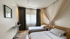 Ett hotellrum med två separata sängar, ett litet nattduksbord, en stol, ett skrivbord med en stol, ett fönster med gardiner och en inramad tavla på väggen.