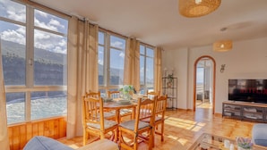 A spacious living room with a large dining table, wooden chairs, and a sofa. There is a TV and a bookshelf in the room.