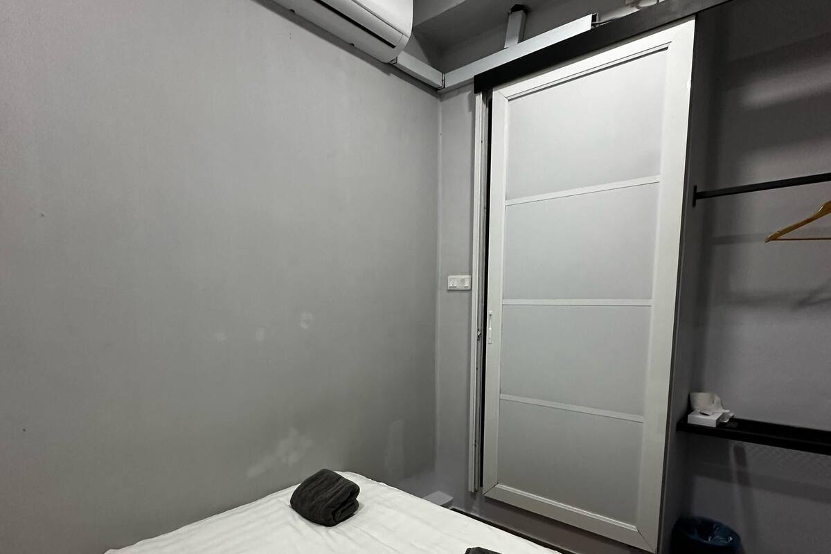 Quarto de hotel com cama, parede branca, unidade de ar condicionado, armário com porta deslizante e uma pequena prateleira com toalha.