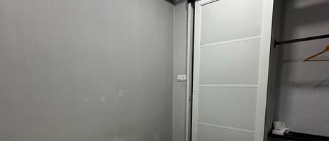 Habitación de hotel con cama, pared blanca, unidad de aire acondicionado, armario con puerta corredera y una estantería pequeña con una toalla.