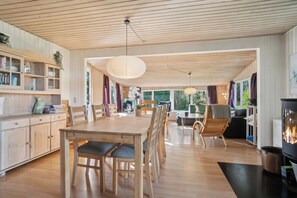 Un vaste espace de vie comprenant une table Ă manger en bois, des chaises, une cheminĂ©e et une grande fenĂȘtre laissant entrer la lumiĂšre naturelle.
