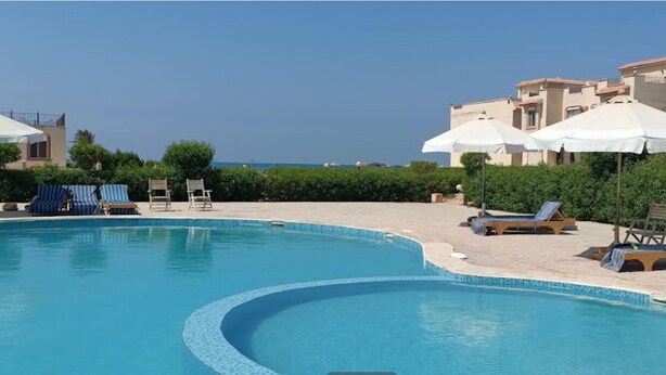 Une piscine entourée de bâtiments donnant sur la mer, avec des chaises longues et des parasols.