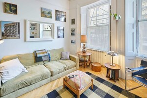 Una sala de estar acogedora con un sofá verde, un otomano estampado, una mesita de madera con una lámpara y un sillón azul.