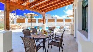 An outdoor patio with a wooden pergola, round dining table, wicker chairs, and umbrellas. - Eliomar Beachside Villa (Chania)