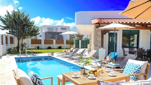 A poolside area with lounge chairs, a table with fruit and drinks, and a view of a modern building. - Eliomar Beachside Villa (Chania)