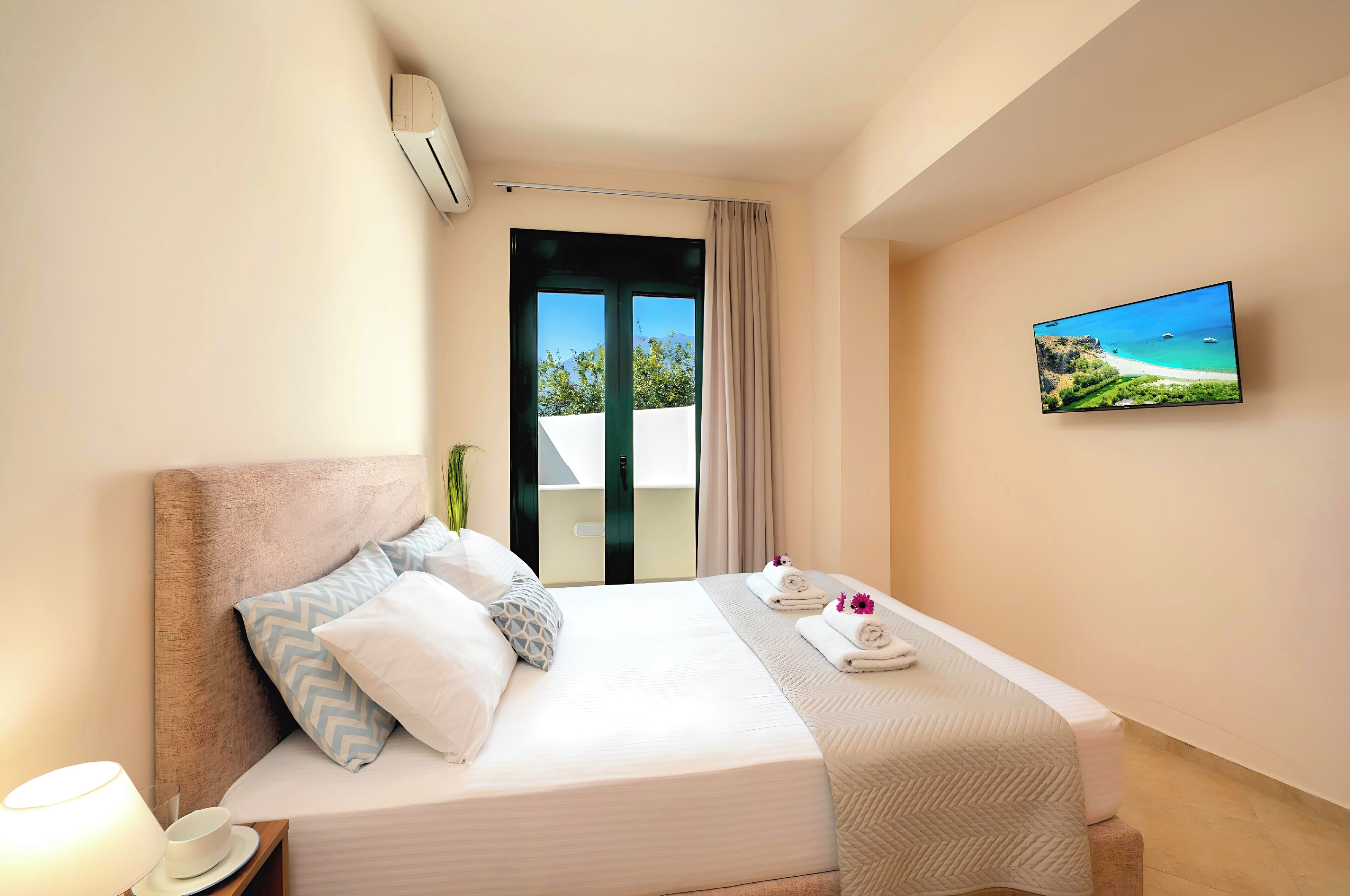 A hotel room with a bed, a TV mounted on the wall, and a view of the outdoors through the window.
