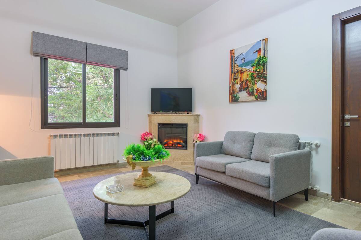 A living room with a grey sofa, a round coffee table with a floral centerpiece, a flat-screen TV above a fireplace, and a painting on the wall.