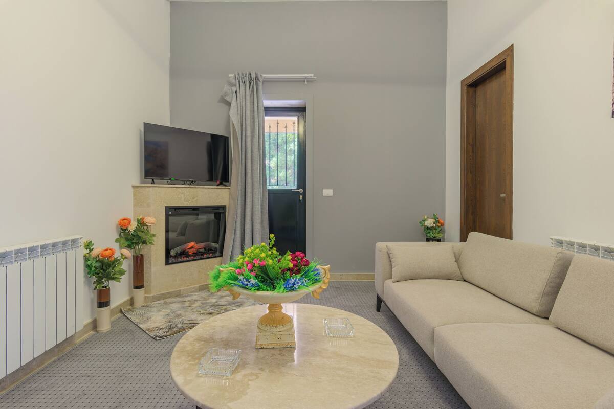 A living room with a beige sofa, a round coffee table with a floral centerpiece, a flat-screen TV on a stand, a fireplace, and a door with a window.