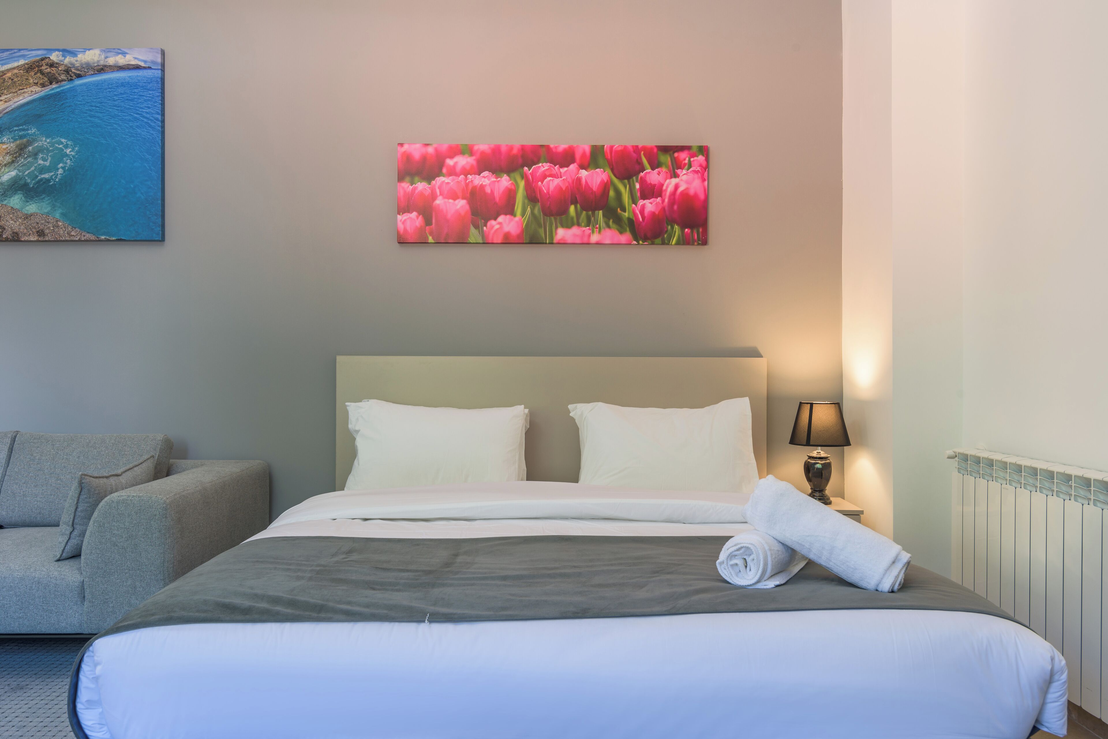 A hotel room with a bed, a grey sofa, a painting of pink tulips, and a lamp.