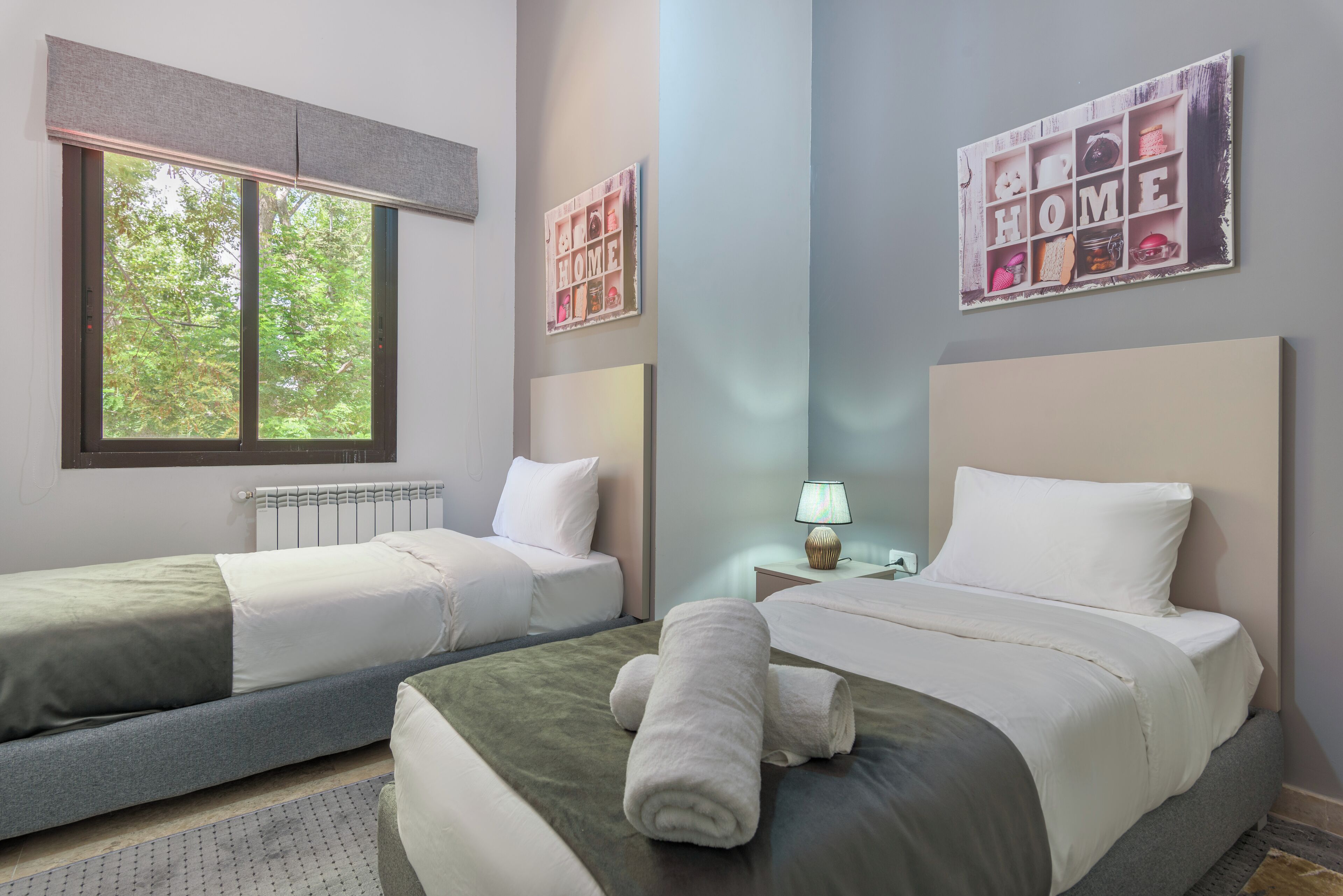 A hotel room with two beds, a window with a view of trees, a wall with a 'HOME' sign, and a lamp on the nightstand.