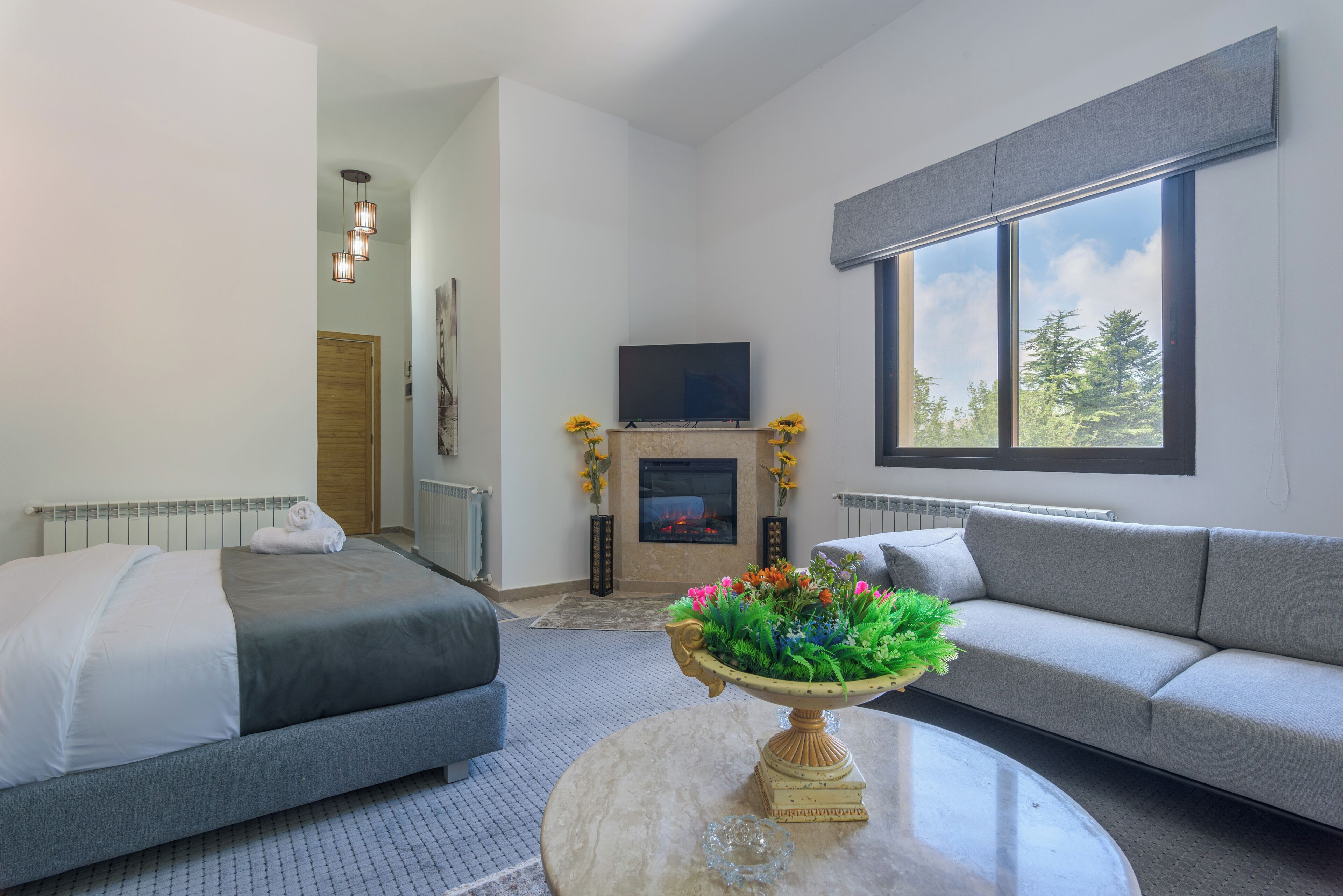 Quarto de hotel moderno com cama, sofá, televisão, lareira e um arranjo de flores sobre uma mesa.