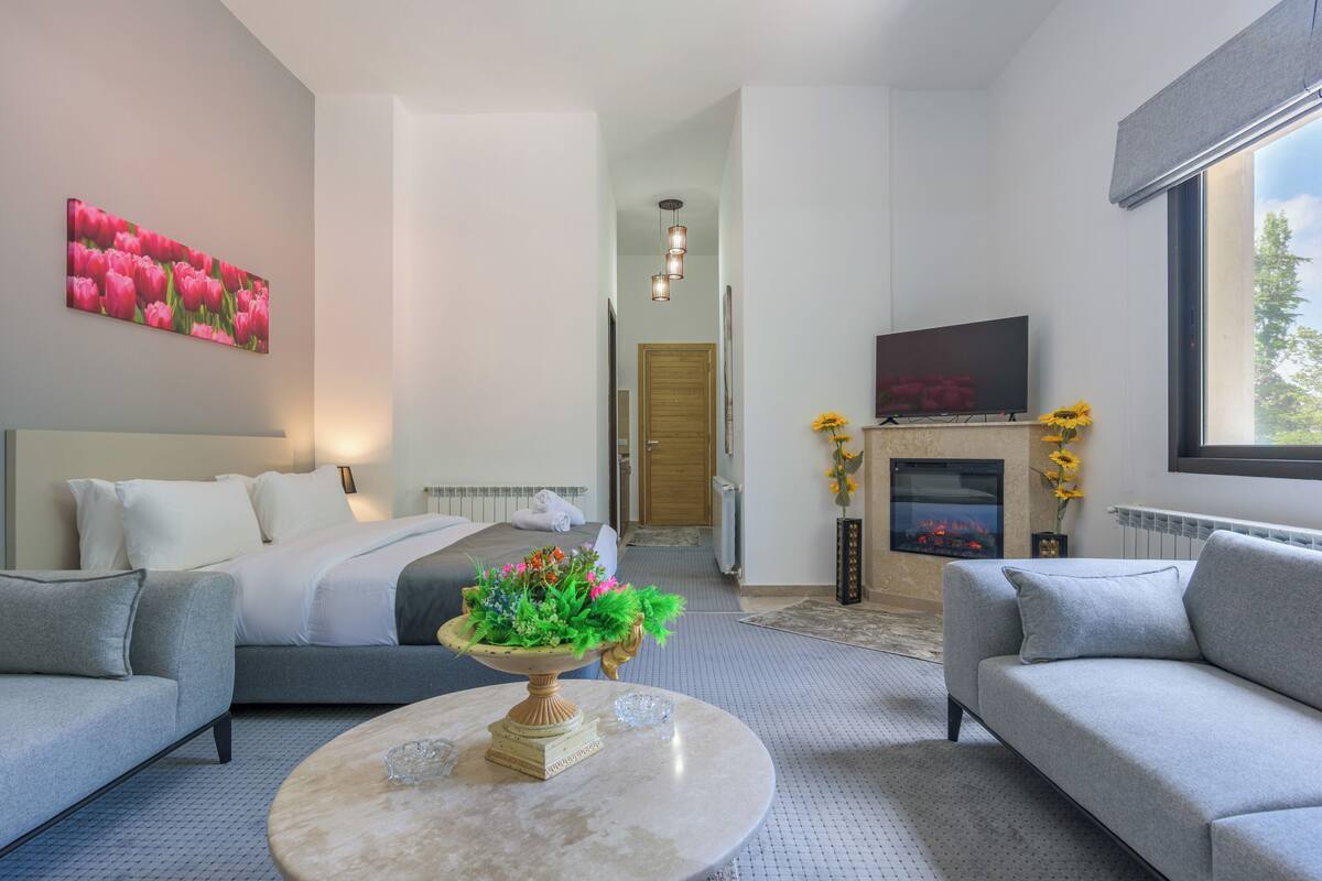 A modern hotel room with a bed, two sofas, a coffee table with a floral centerpiece, a TV, a fireplace, and a large window.