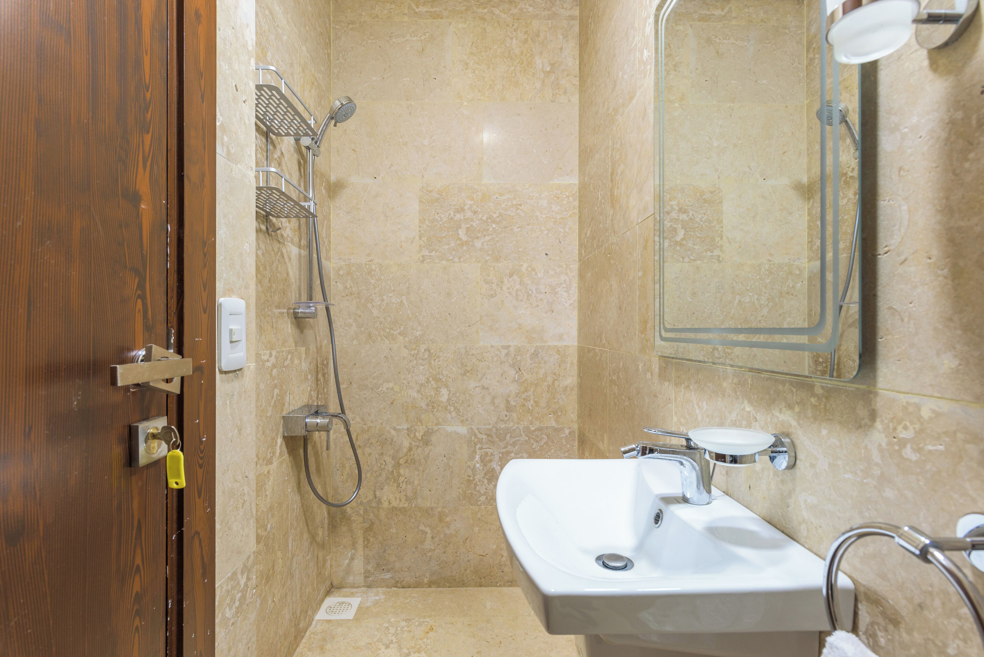 Ein Badezimmer mit Holztür, einer Dusche mit Metallablage, einem Waschbecken mit Spiegel darüber und beige gefliesten Wänden.