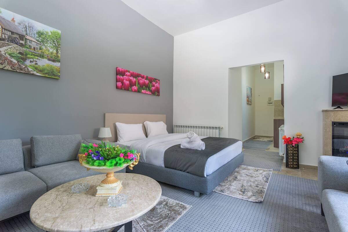 A modern hotel room with a bed, sofa, and a dining table with a floral centerpiece.