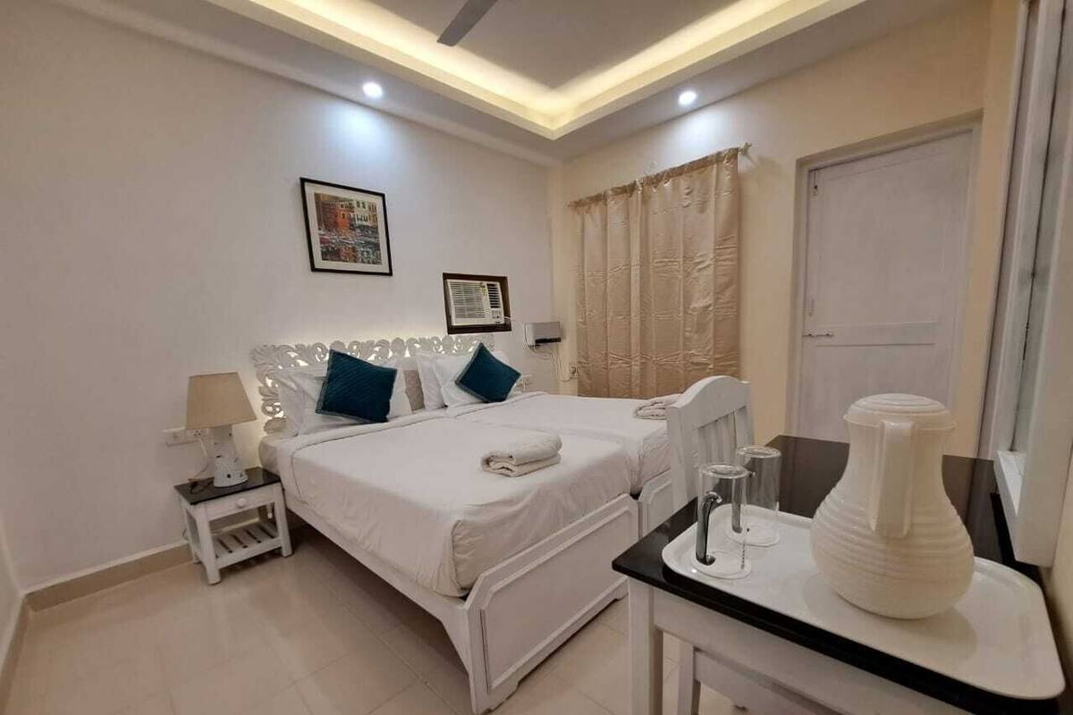 A hotel room with a bed, bedside table, lamp, air conditioning unit, and a small table with a vase.